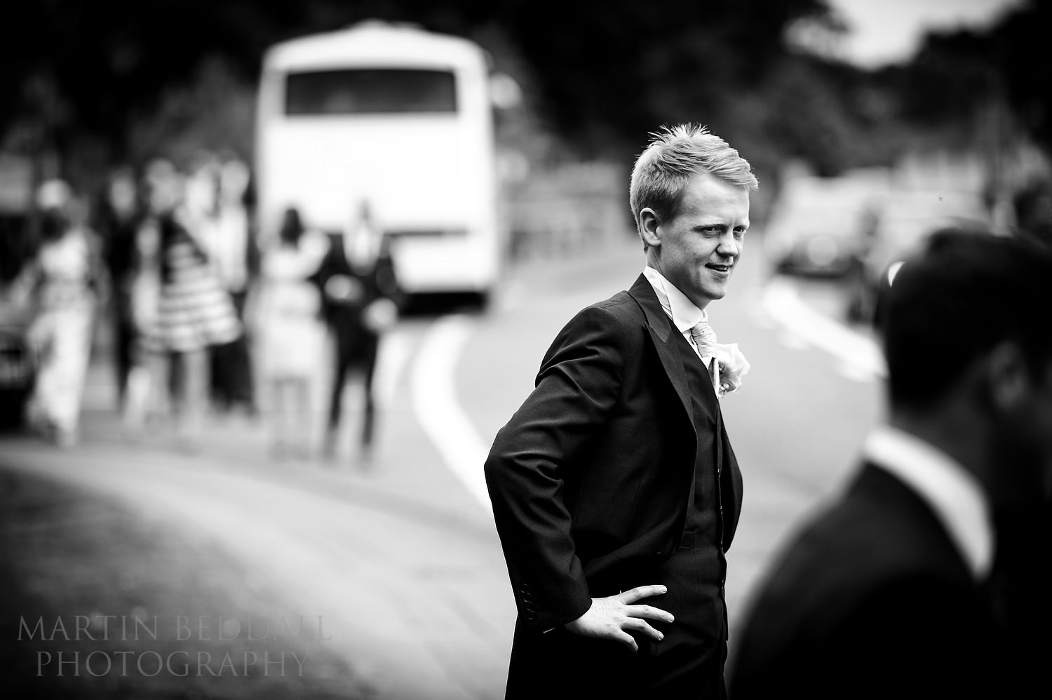Groom outside Chiddingfold church as guests arrive by coach