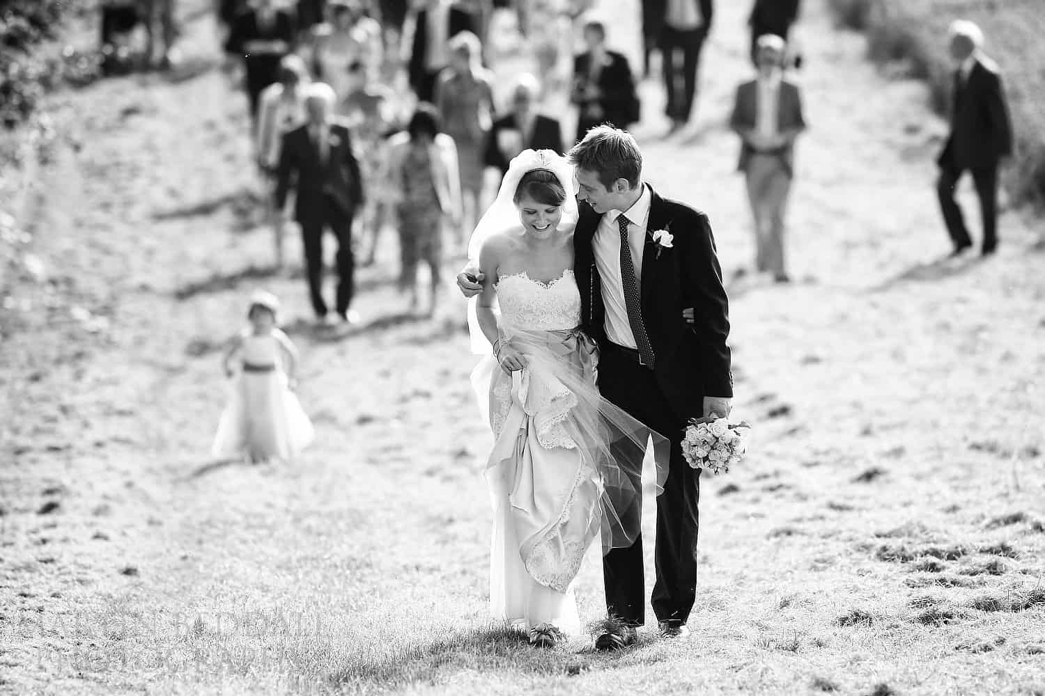 Bride and groom lead the wedding guests across sunny fields to the wedding reception